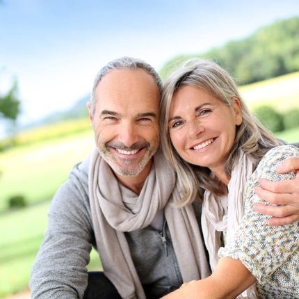 Smiling middle-aged couple outdoors in a bright, natural setting, embracing affectionately with a blurred green landscape in the background.
