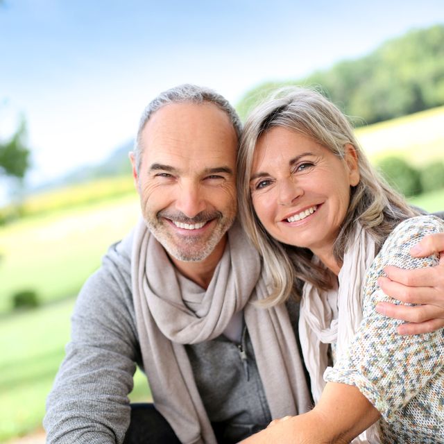 Smiling middle-aged couple outdoors in a bright, natural setting, embracing affectionately with a blurred green landscape in the background.