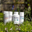 Several white supplement containers with various labels including multivitamin, sleep support, and daylight/nighttime formulas, placed on green grass with sunlight and blurred natural background.