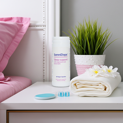 White bedside table with a bottle of SomniDopa sleep support capsules, two loose capsules, a folded white towel with white flowers, and a potted green plant, next to a pink bed.
